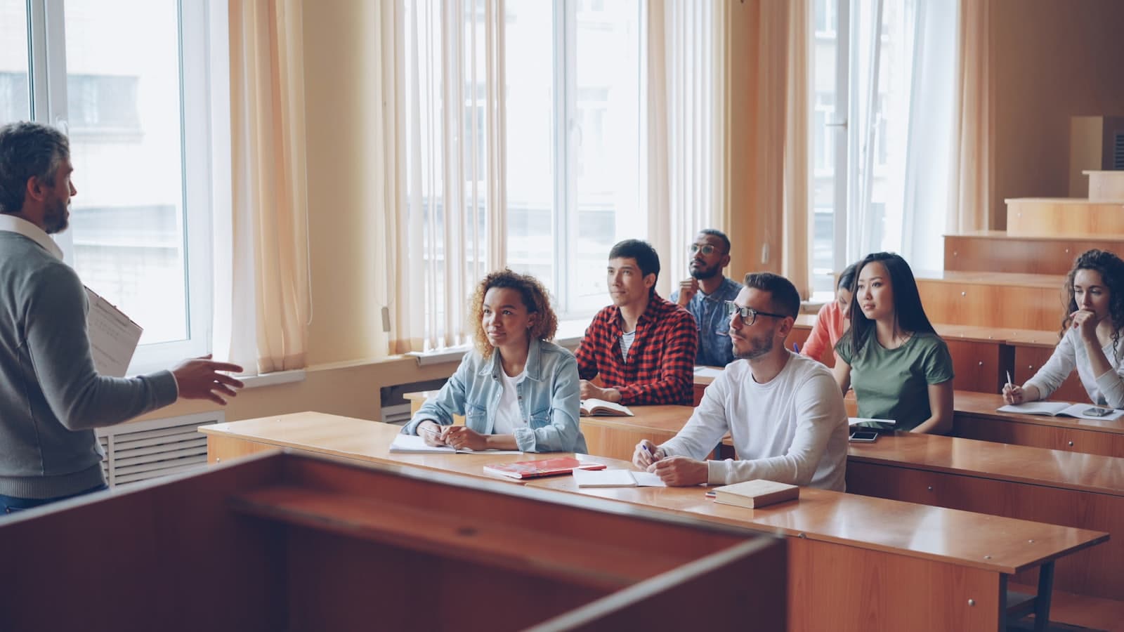 Students at tiered desks in a lecture room, listening and taking notes while an instructor speaks