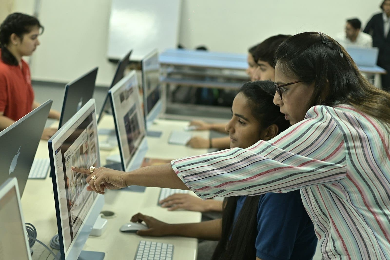 An instructor leaning in to point at a student's screen during a lab session, other students working along the same bench